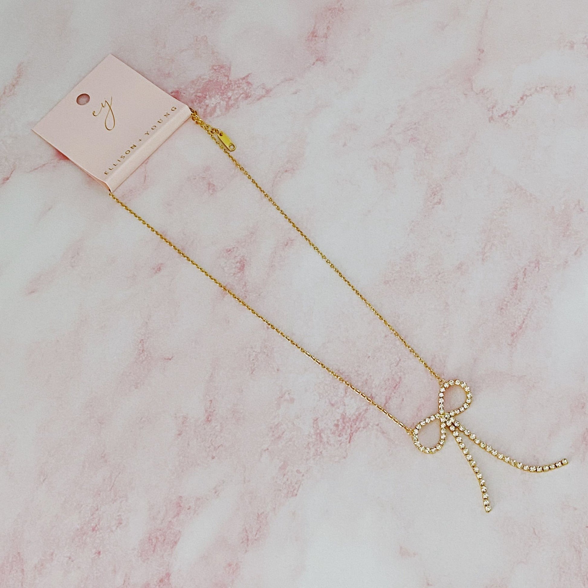 Gold necklace with a sparkling rhinestone bow pendant, elegantly displayed on a pale pink marble background.