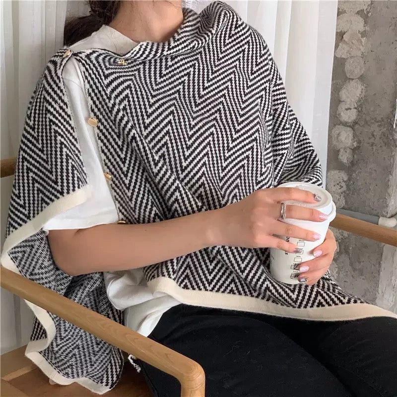 Woman wearing a stylish black and white herringbone patterned poncho, sitting in a cozy setting while holding a coffee cup, showcasing comfort and modern design.