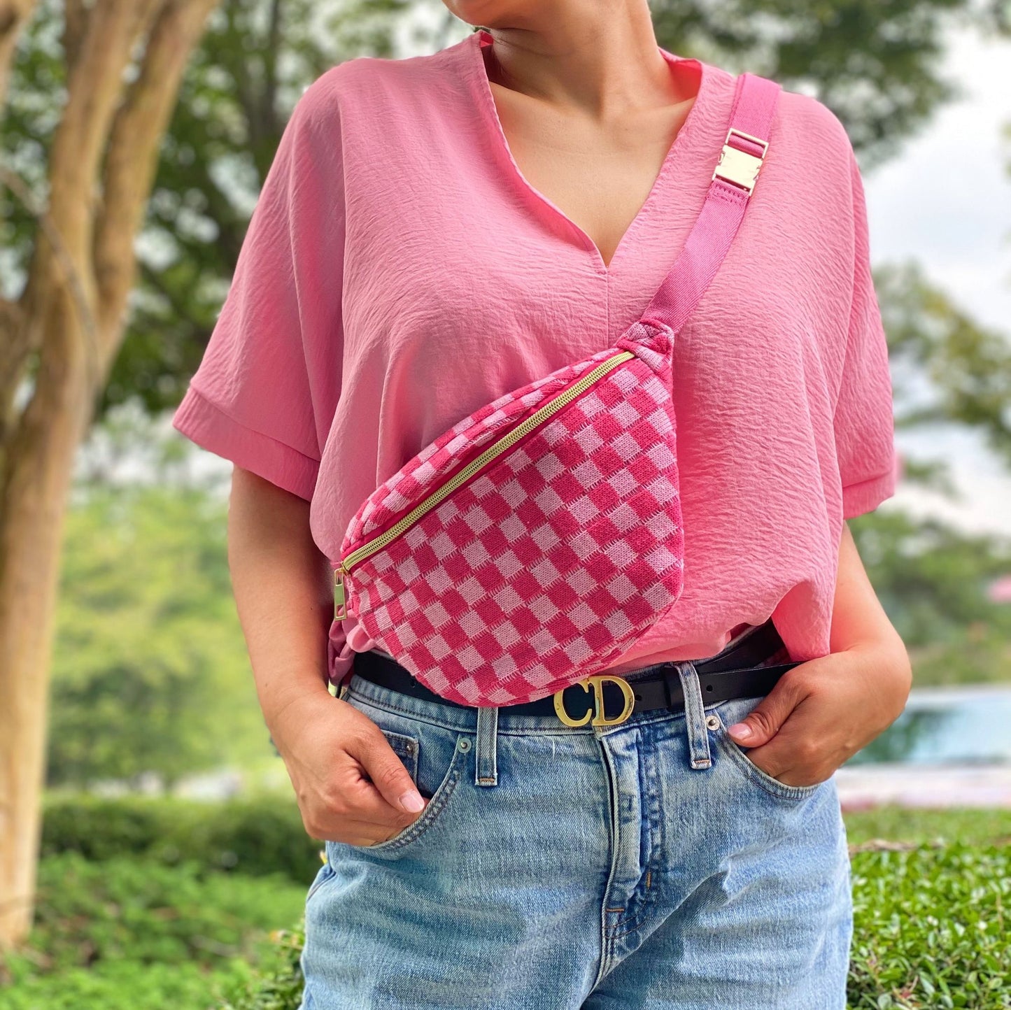Woman wearing a pink oversized top and light blue jeans, showcasing a stylish pink gingham fanny pack with a gold zipper, set against a lush outdoor background.