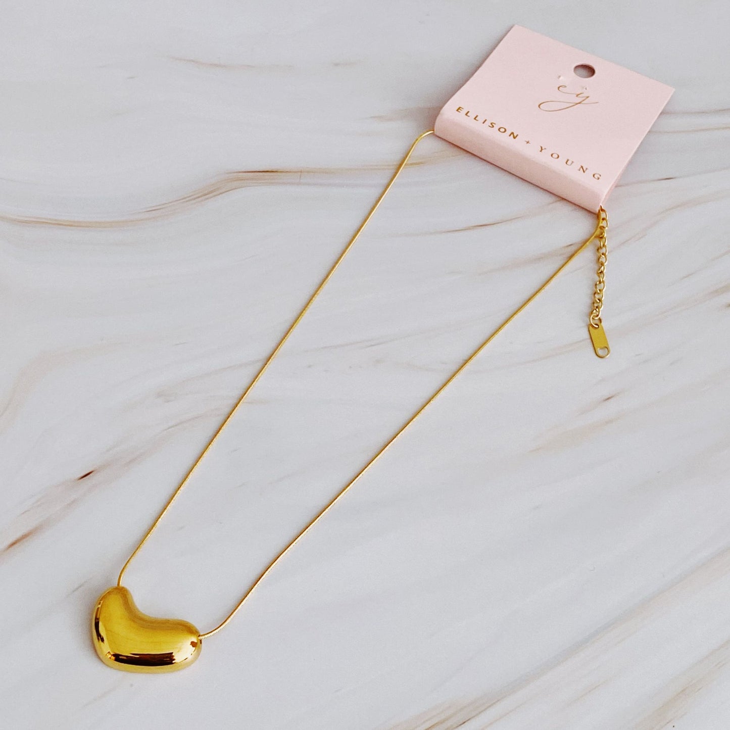 Gold heart-shaped pendant necklace displayed on a marble surface with a pink tag reading "Ellison + Young".