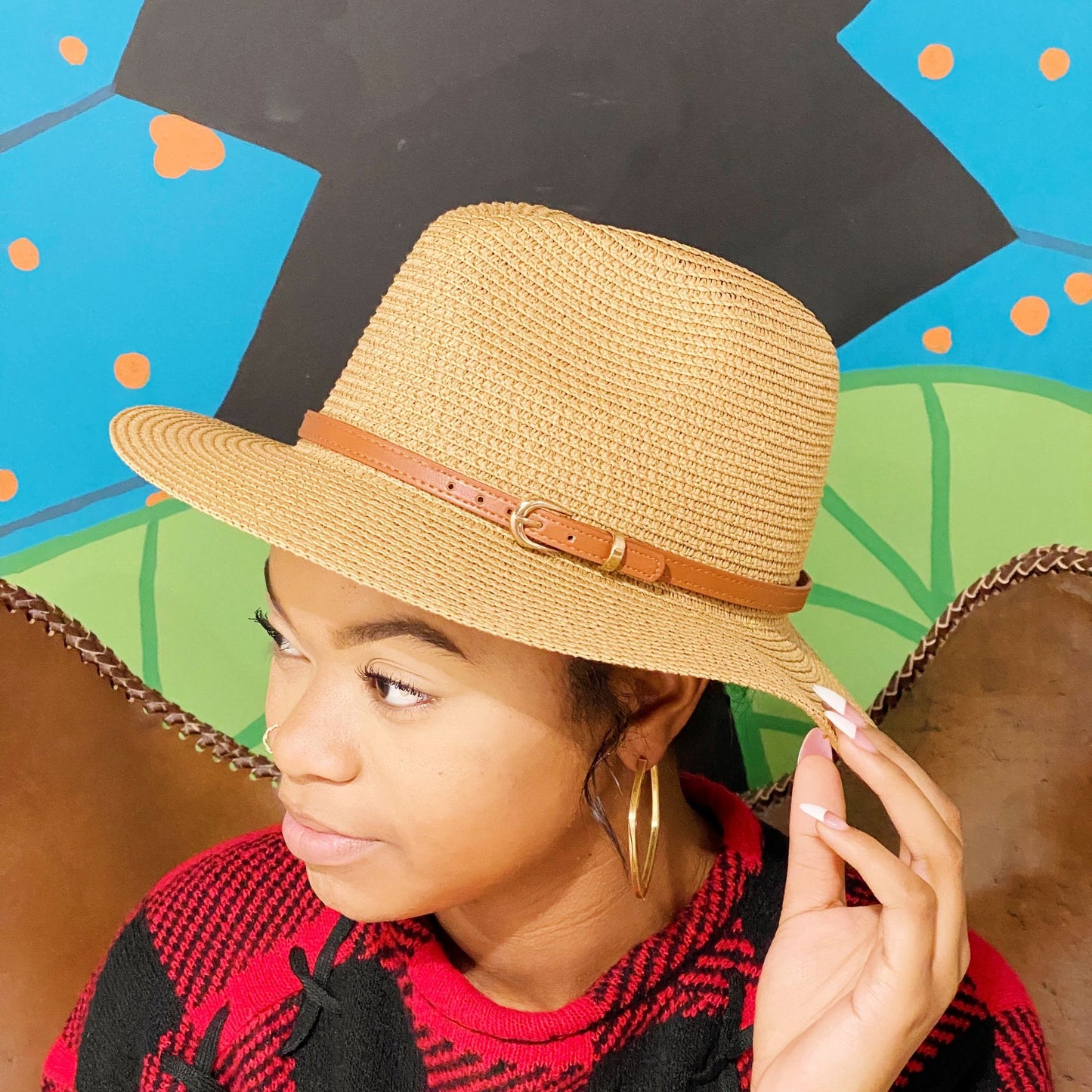 Stylish straw fedora hat with a brown leather band, worn by a person with curly hair, against a colorful mural background.
