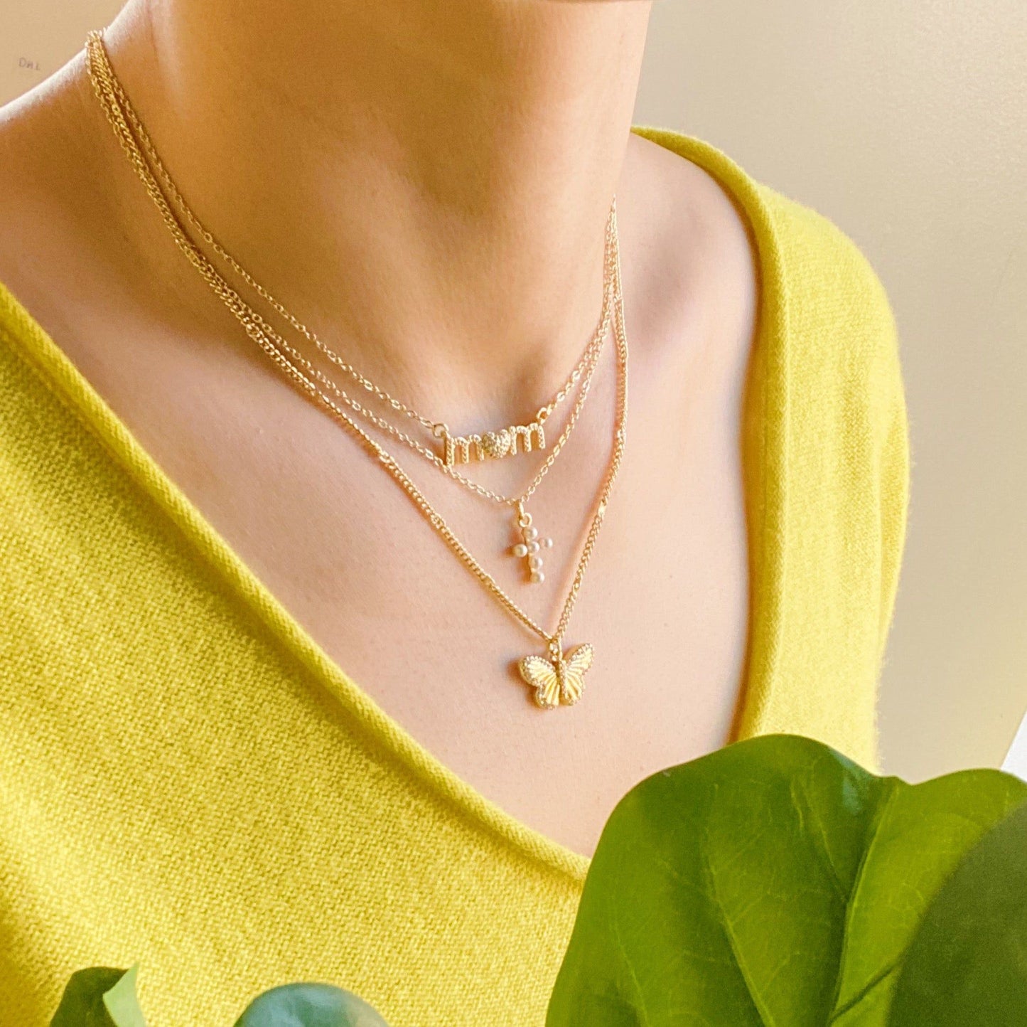 Layered gold necklaces featuring a 'mom' pendant, pearl accents, and a butterfly charm, worn by a model in a bright yellow sweater, complemented by green foliage.