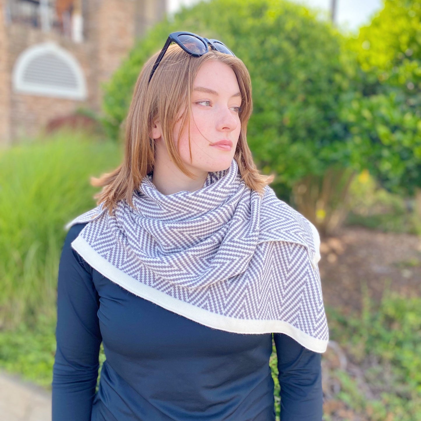 Woman wearing a stylish gray and white herringbone-patterned scarf over a black long-sleeve shirt, set against a lush green background.