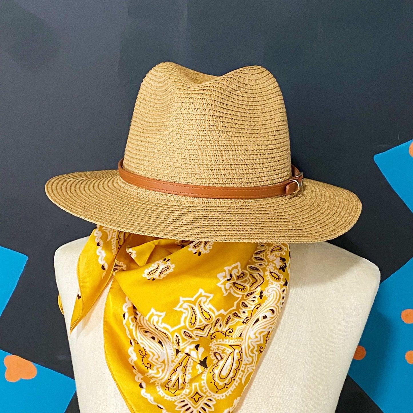 Straw wide-brimmed hat with a brown leather band displayed on a mannequin, paired with a bright yellow paisley bandana, against a colorful background.