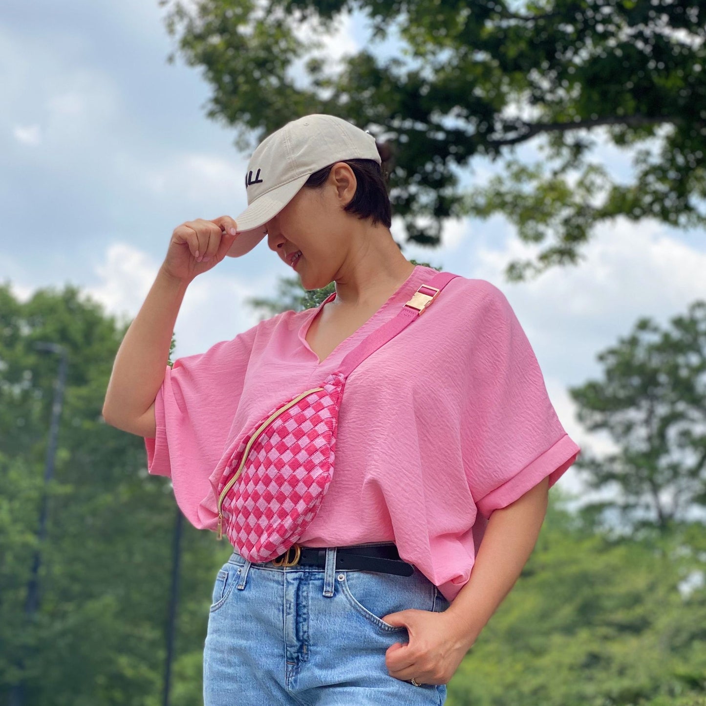 A woman wearing a vibrant pink oversized top and light blue denim shorts, posing outdoors with a trendy pink checkered crossbody bag, against a backdrop of green trees and a cloudy sky.