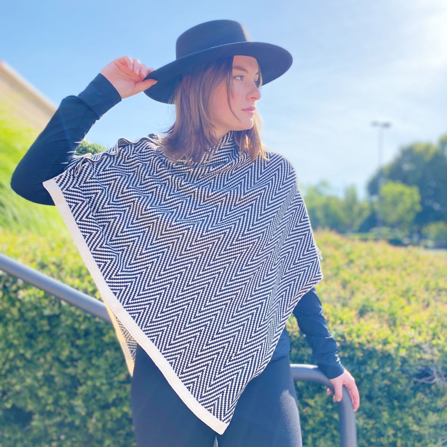 Fashionable black and white herringbone poncho draped elegantly over a model's shoulder, complemented by a stylish black wide-brim hat against a bright outdoor background.