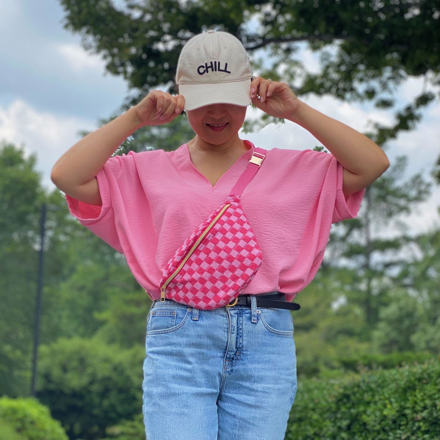 A person wearing a stylish pink oversized shirt and light blue jeans, accessorized with a vibrant pink checkered fanny pack, positioned against a lush green outdoor background.