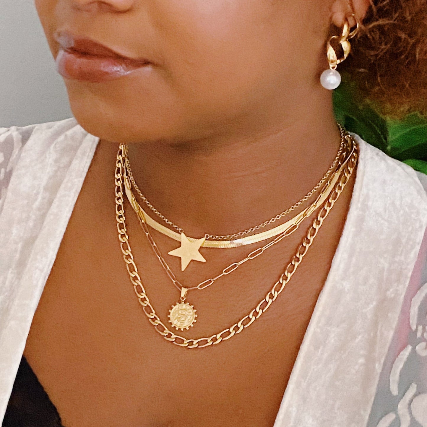 Close-up image of a woman's neck adorned with a layered gold necklace featuring a star pendant and a sun charm, complemented by a pearl hoop earring, showcasing elegant gold jewelry and a stylish look.