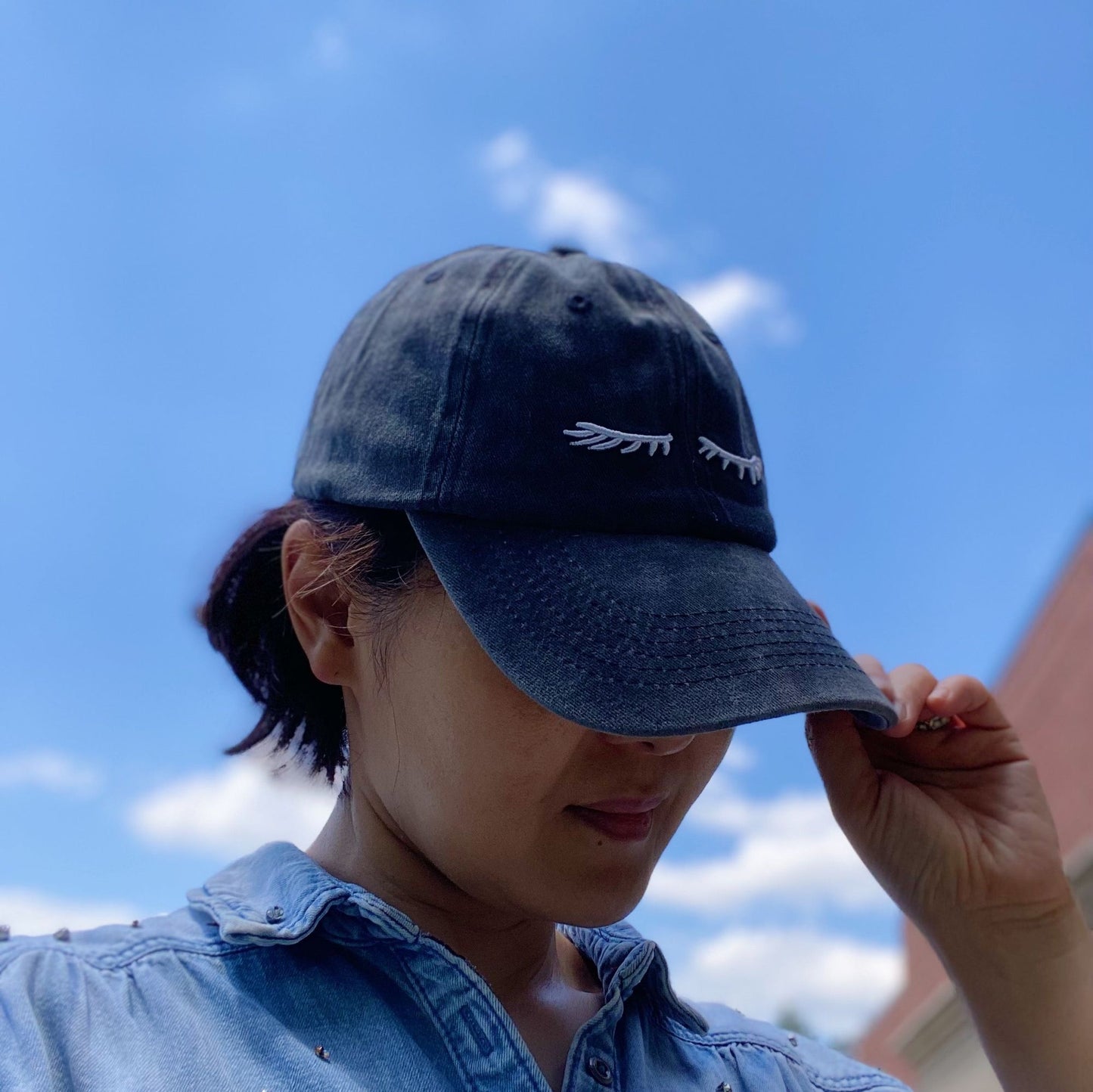 Stylish navy blue baseball cap with white embroidered leaf design, worn with casual denim shirt against a bright blue sky background.