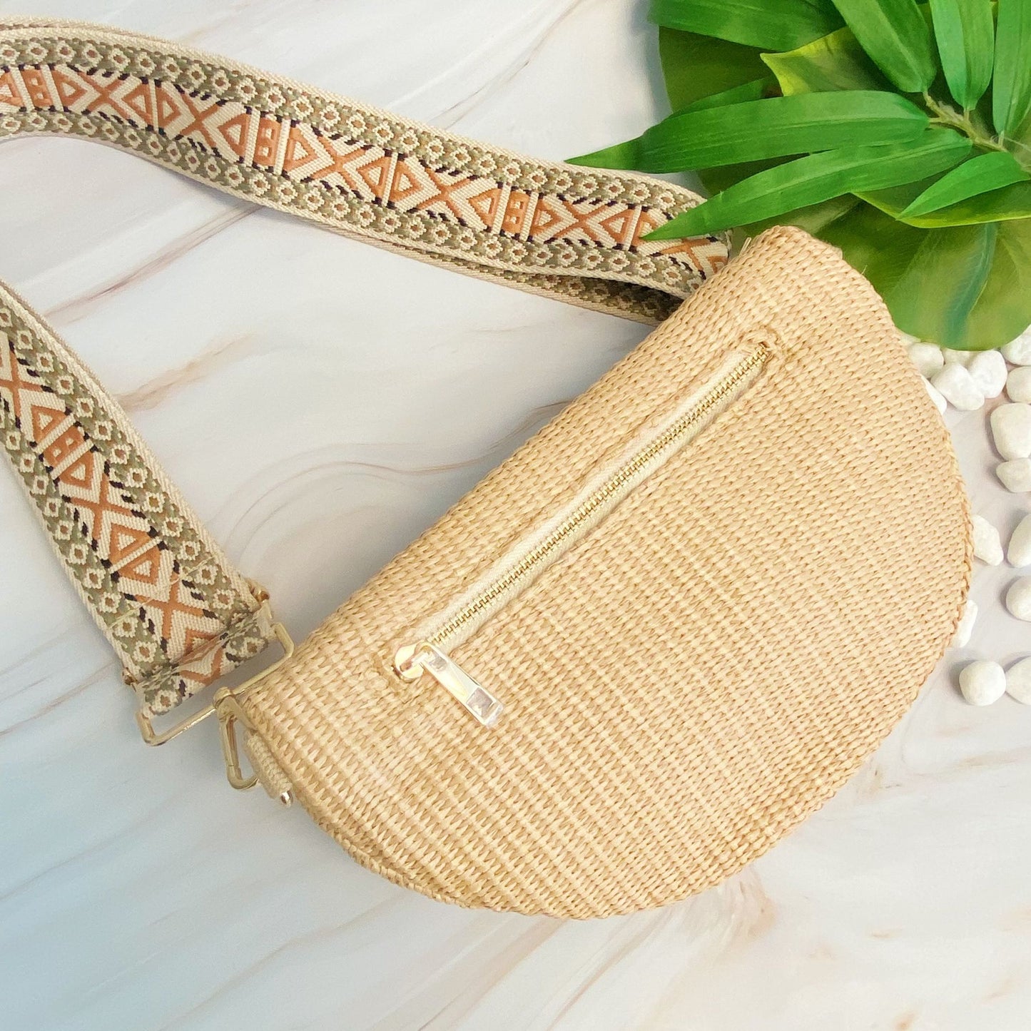 Woven straw handbag with a gold zipper and decorative patterned strap, set against a light background with tropical leaves and pebbles.