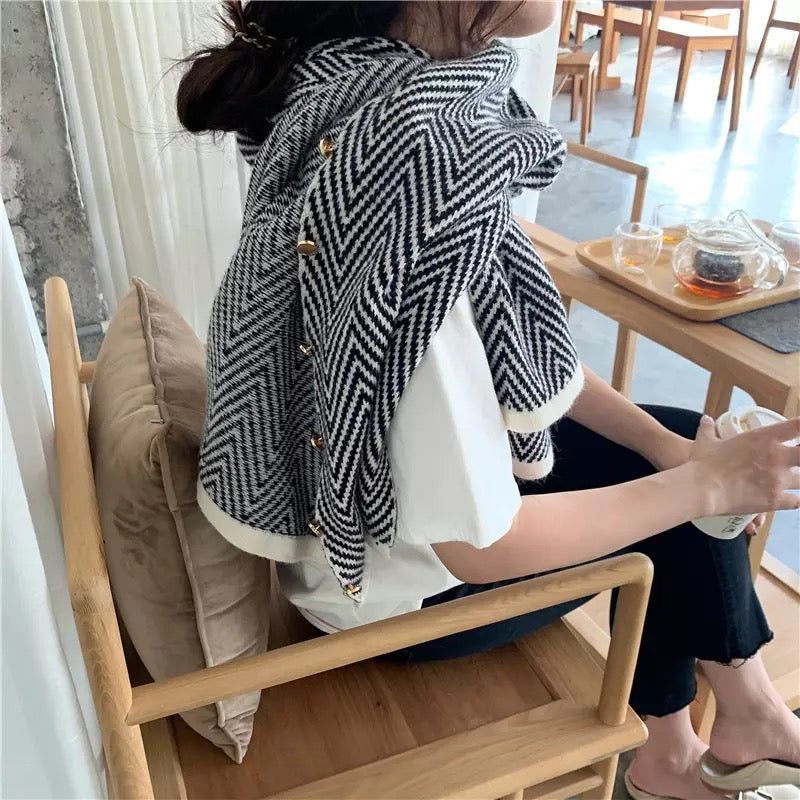 Elegant black and white herringbone-patterned scarf draped over a woman's shoulders, featuring gold button accents, as she sits comfortably in a modern cafe setting.