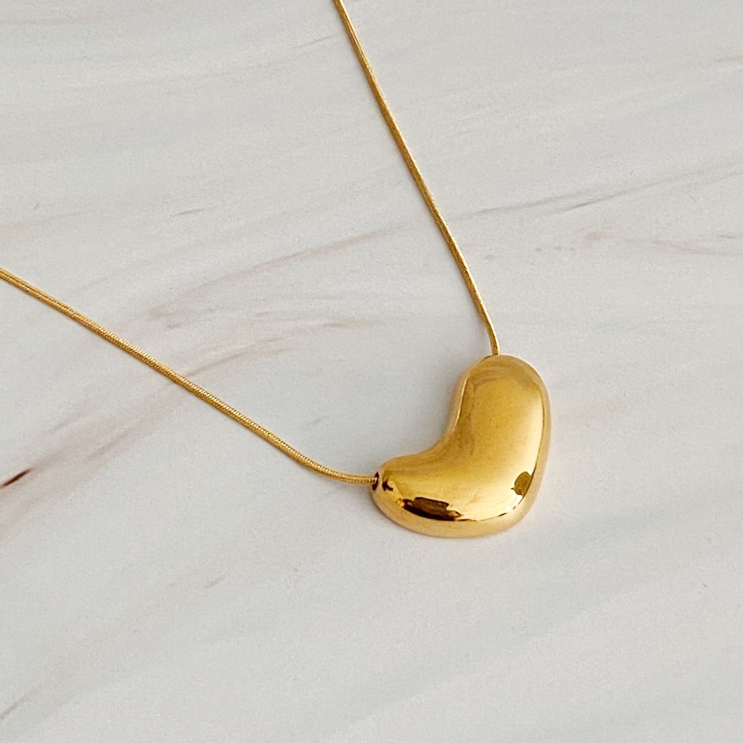 Stylish gold heart-shaped pendant necklace on a delicate chain, resting on a light marble surface.