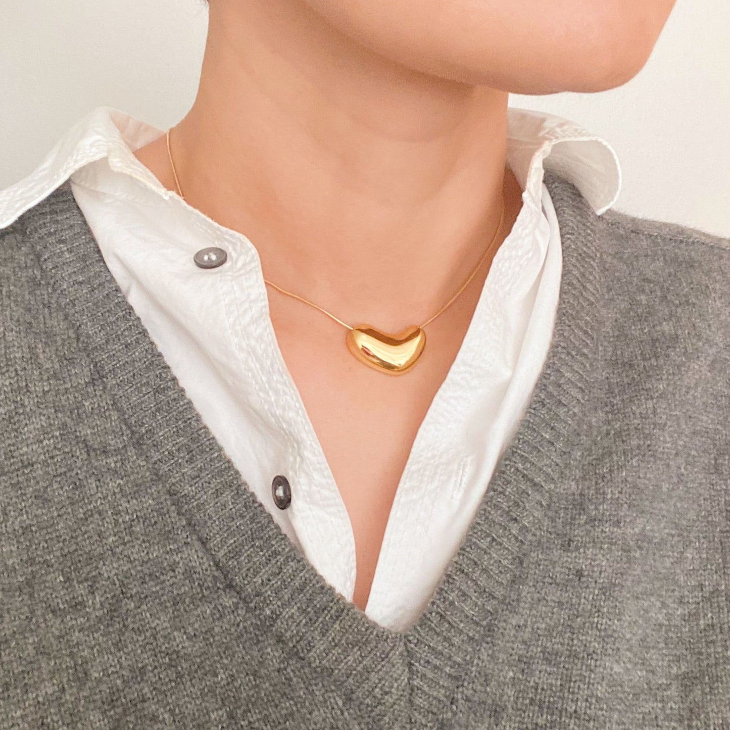 Stylish gold heart-shaped pendant necklace worn over a white button-up shirt, paired with a grey sweater, showcasing contemporary jewelry fashion.