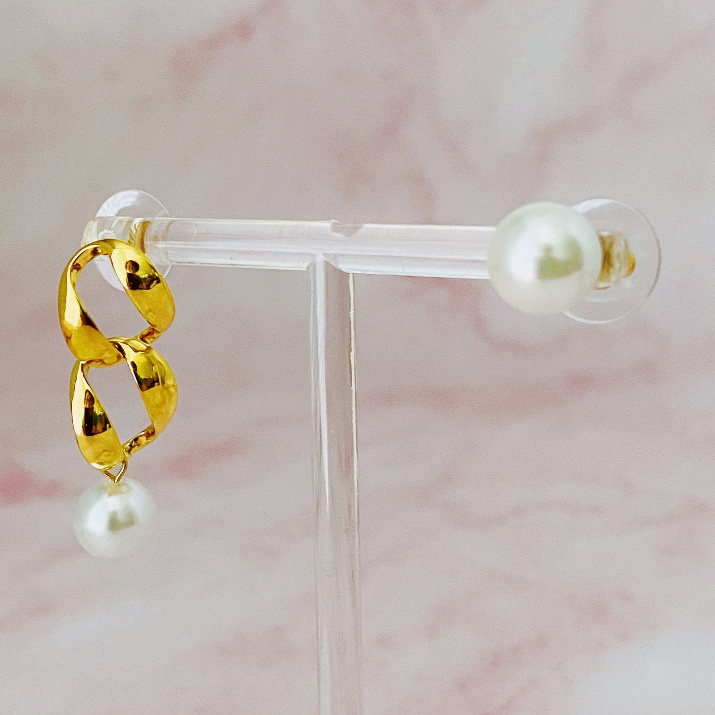 Stylish gold chain-link earring with a pearl drop, displayed on a clear acrylic stand against a soft pink background.
