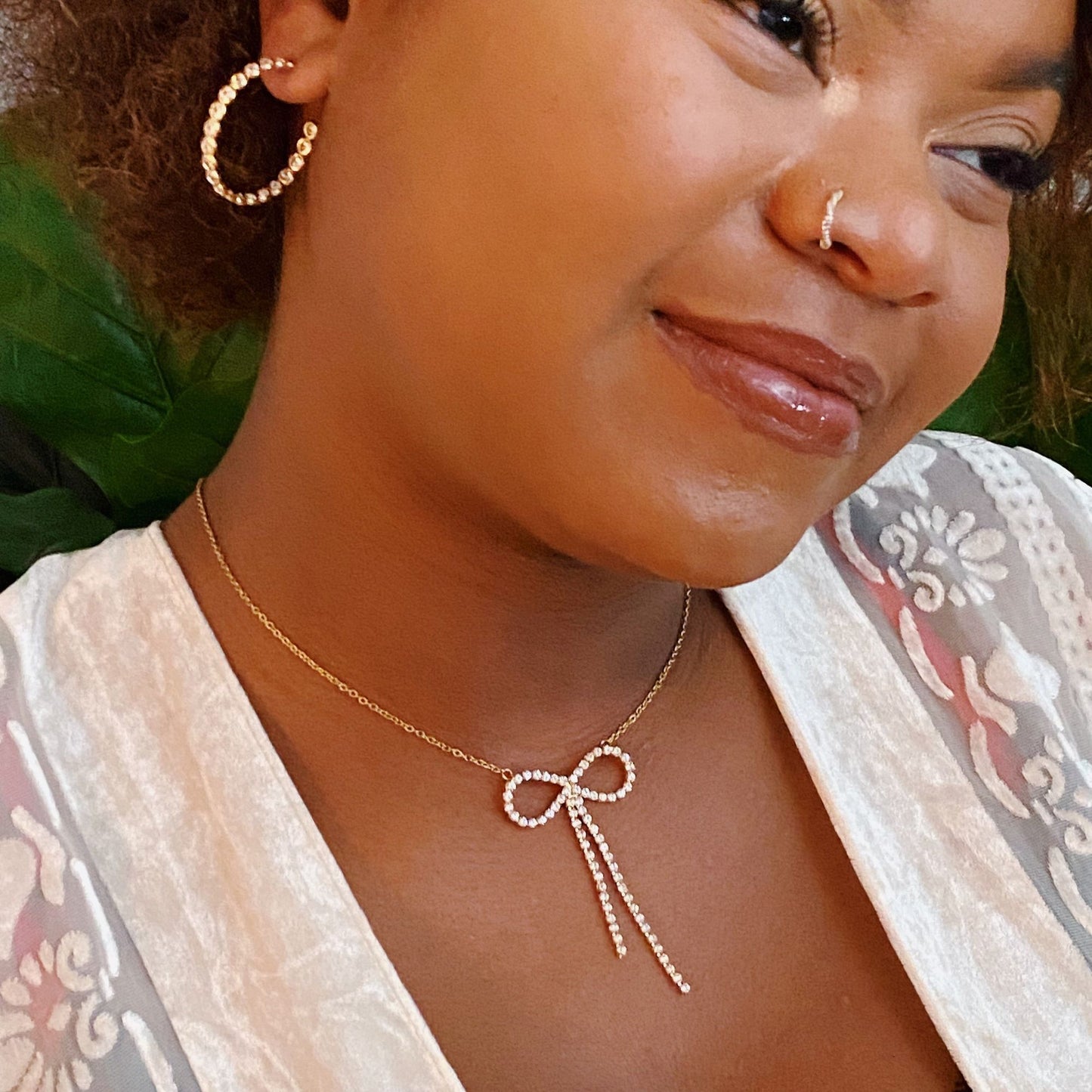 Gold necklace with sparkling rhinestone bow pendant, complemented by matching hoop earrings, against a backdrop of lush greenery.