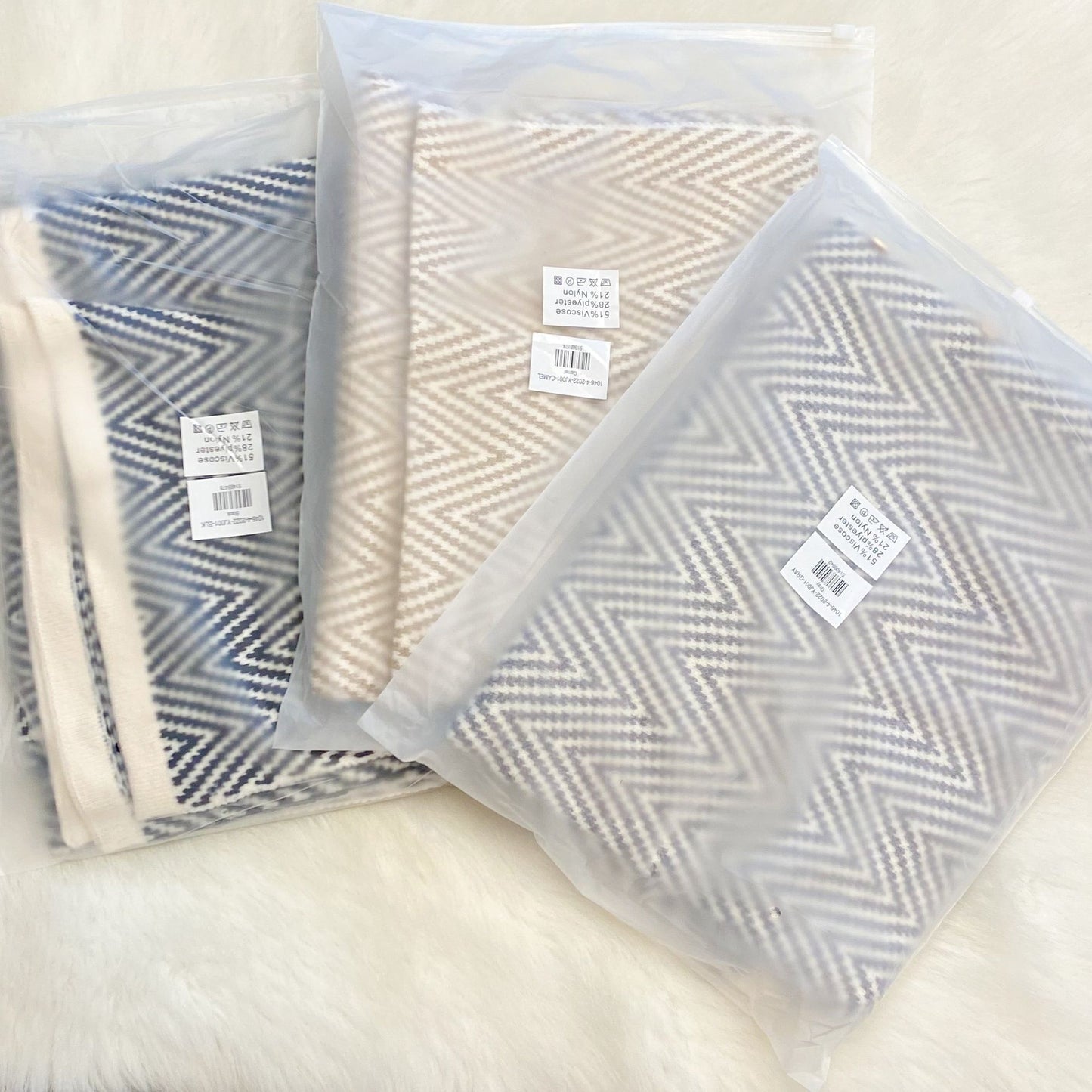 Three herringbone patterned blankets in protective packaging, featuring colors of blue, beige, and light gray, arranged on a soft white surface.
