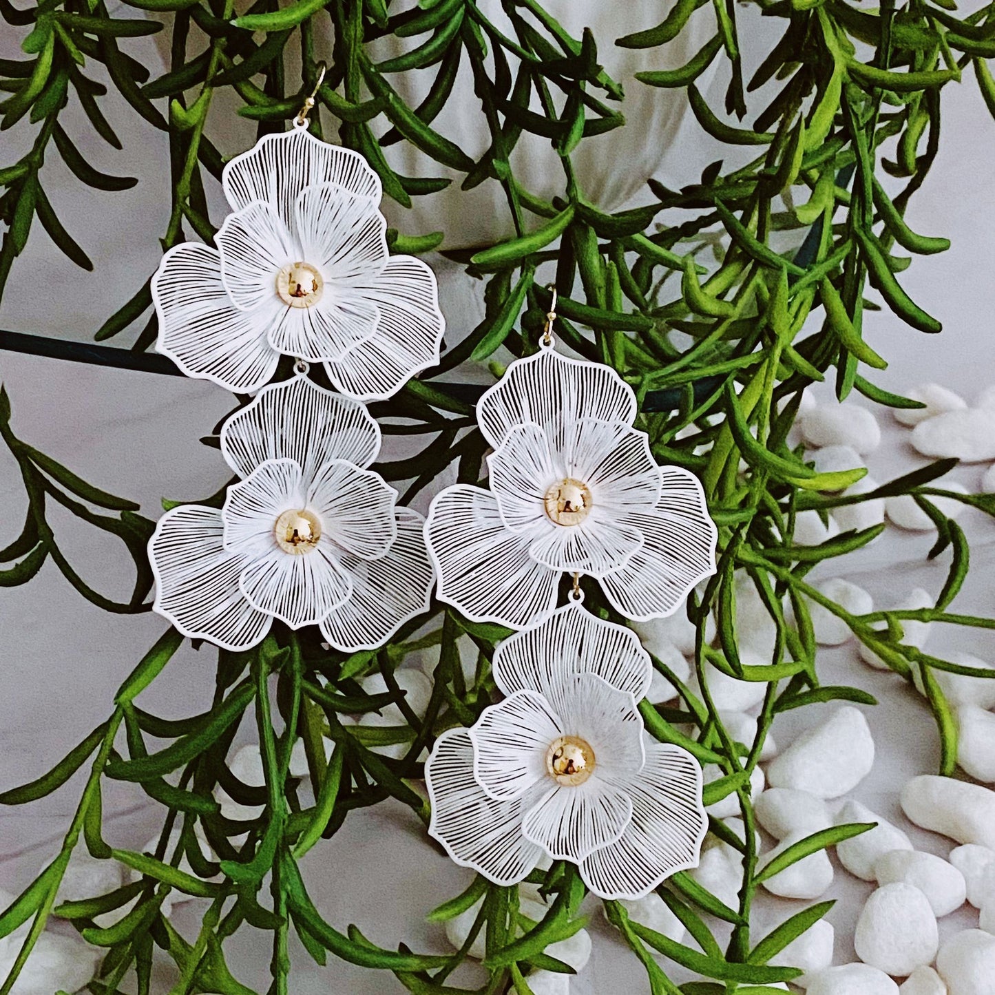 Elegant white floral earrings featuring intricate lace-like details and gold center, hanging among lush greenery and decorative pebbles, perfect for adding a touch of sophistication to any outfit.
