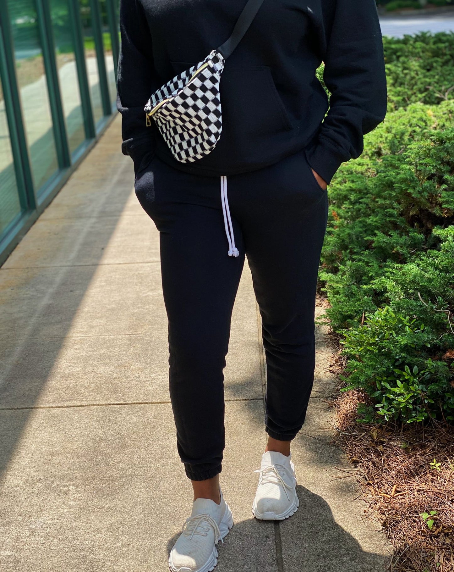 Woman in a black hoodie and matching jogger pants, wearing white sneakers, with a stylish black and white checkered crossbody bag, standing on a sunny sidewalk with greenery in the background.