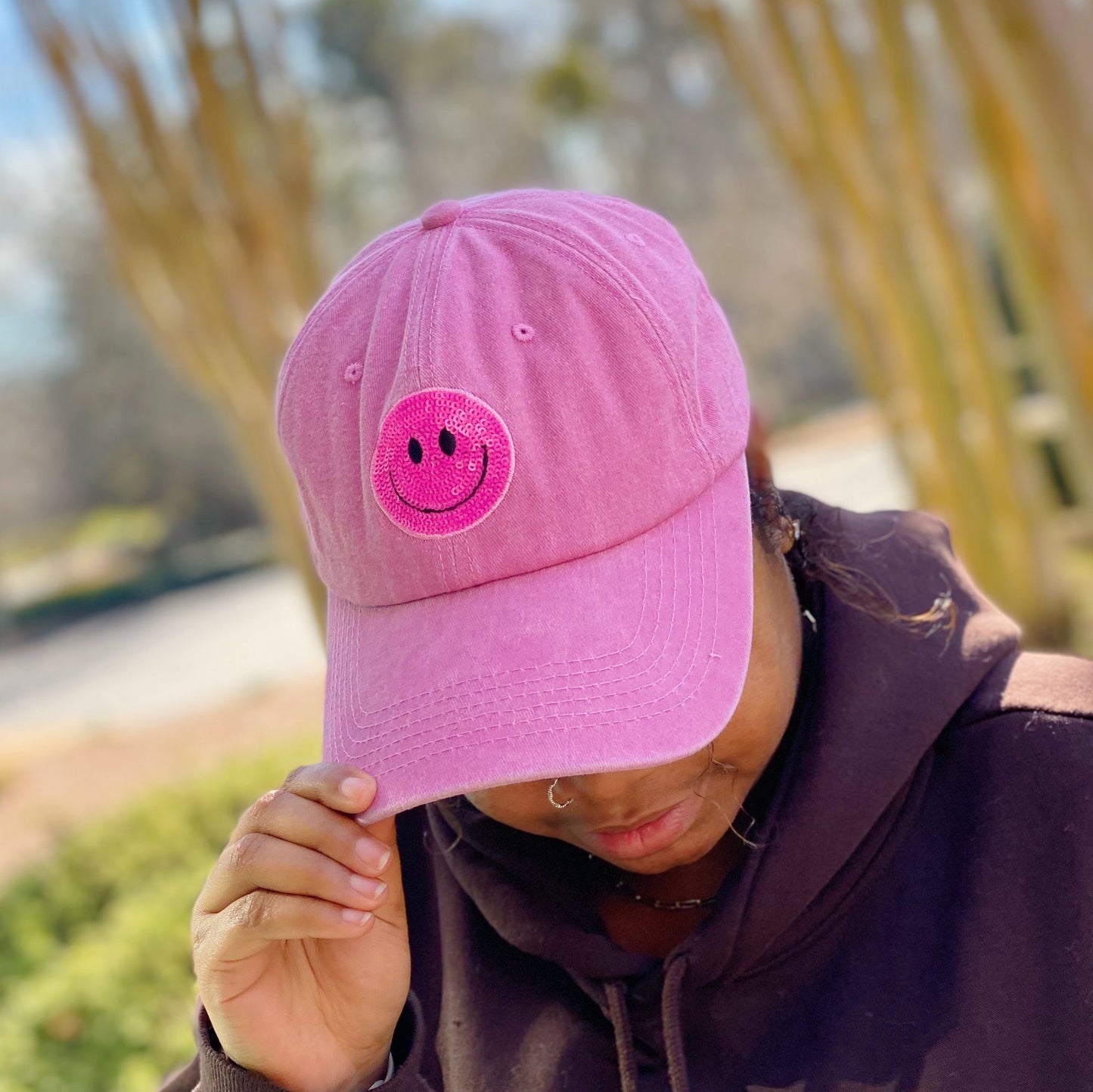A woman in a brown hoodie wears a stylish pink cap with a smiling face patch, captured outdoors with greenery in the background.