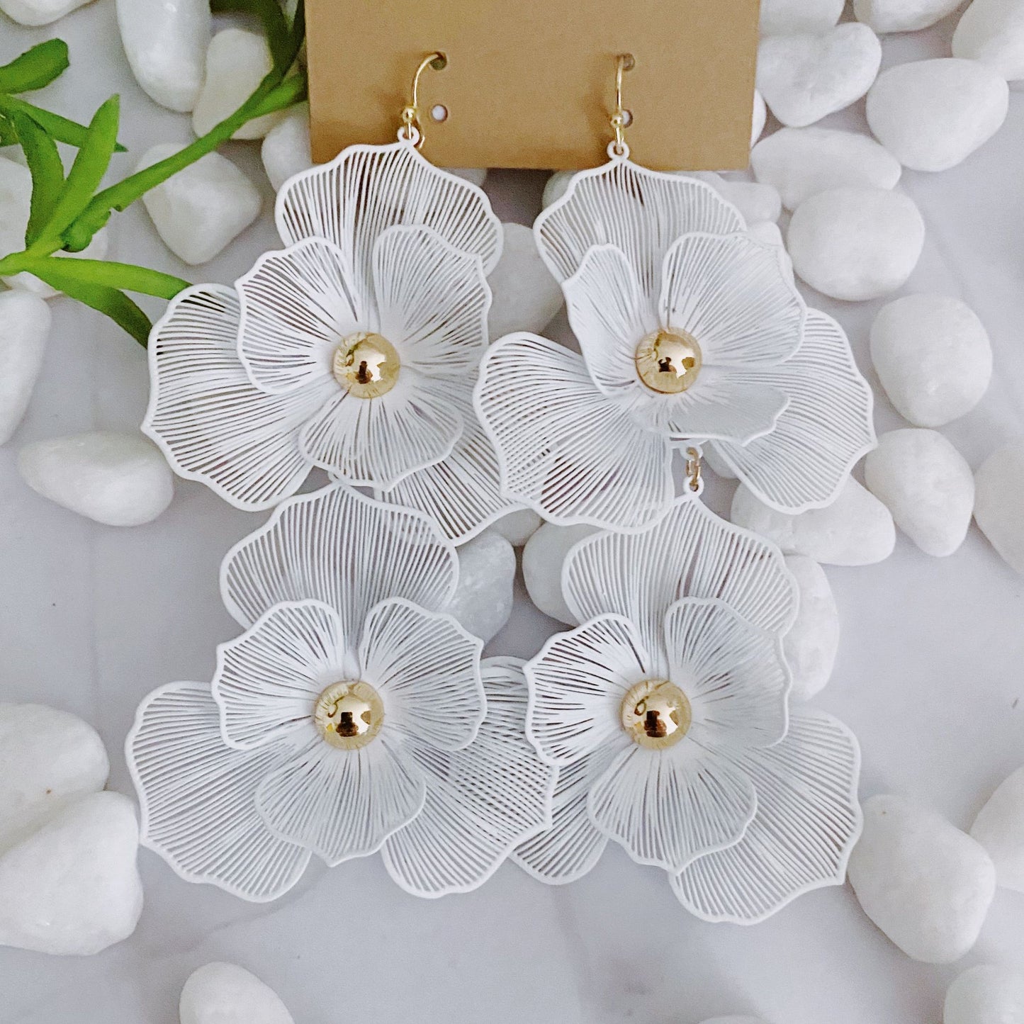 Elegant white floral earrings with gold accents displayed on a neutral background surrounded by smooth white stones.