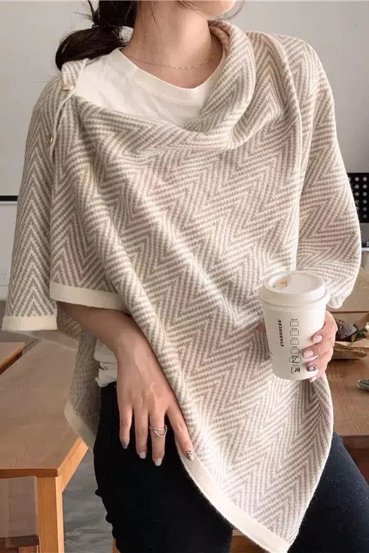 Woman wearing a stylish beige and cream herringbone patterned poncho, holding a coffee cup, showcasing a chic and cozy autumn outfit.