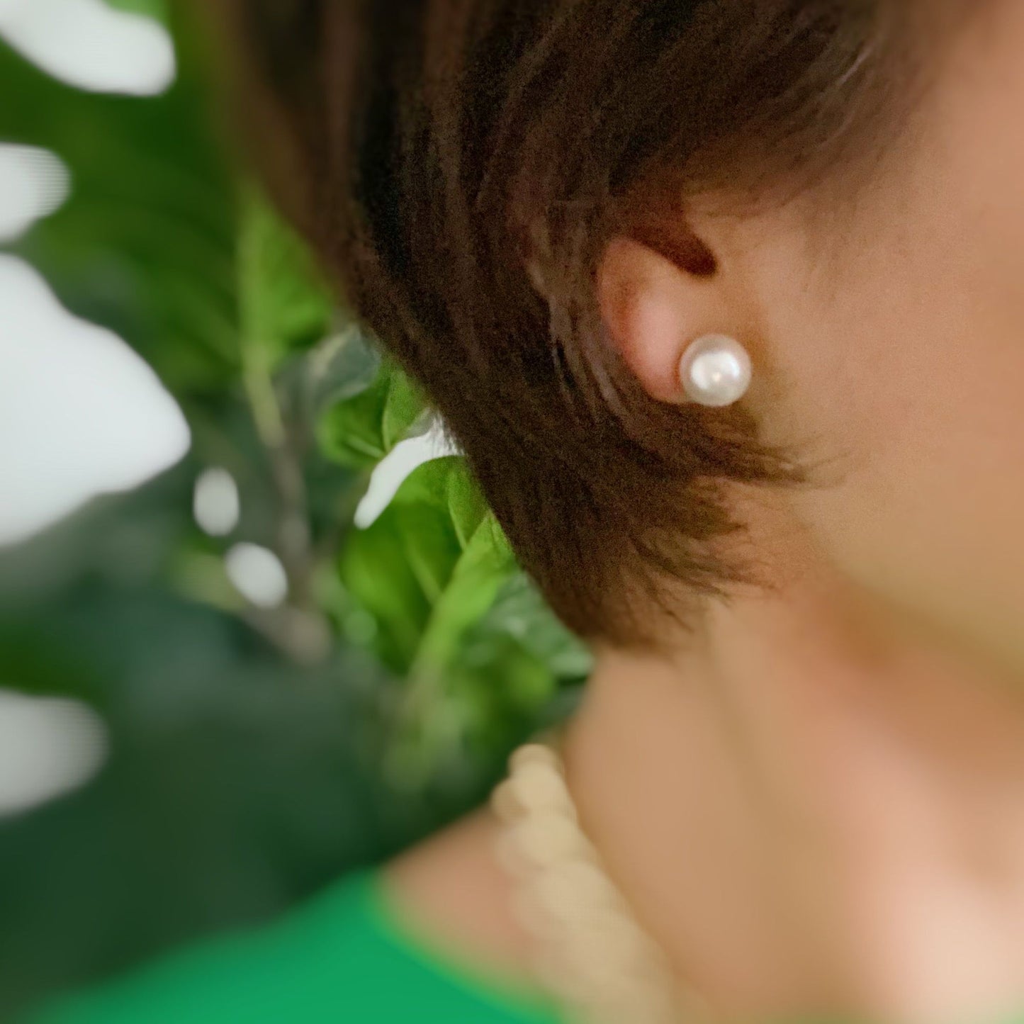 Close-up of a woman wearing a classic pearl stud earring, with her short hair styled elegantly, set against a background of lush green foliage.