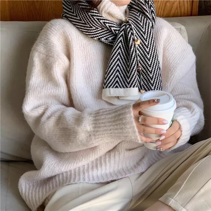 Cozy woman wearing a soft beige oversized sweater and a black and white chevron scarf, holding a coffee cup while relaxing on a couch.