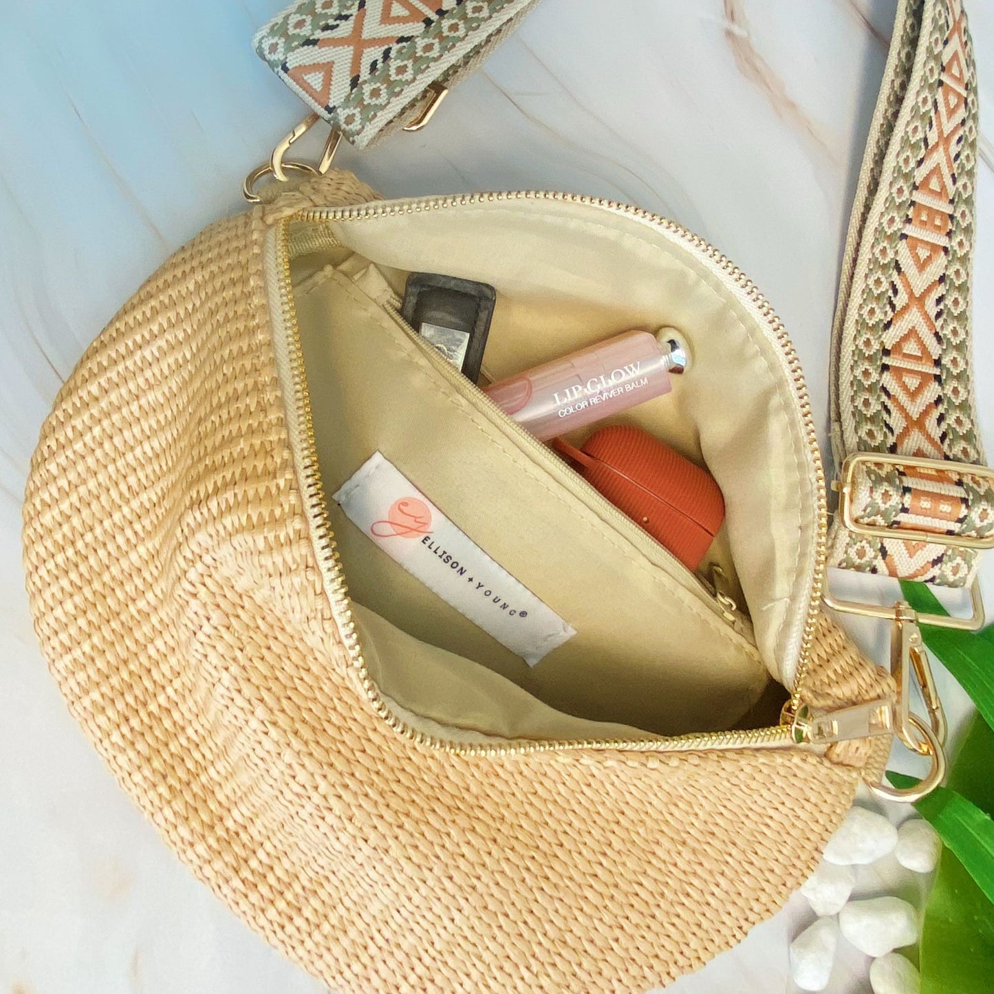 Inside view of a stylish beige woven crossbody bag showcasing its spacious interior with beauty products including a lip balm, all on a light marble background.