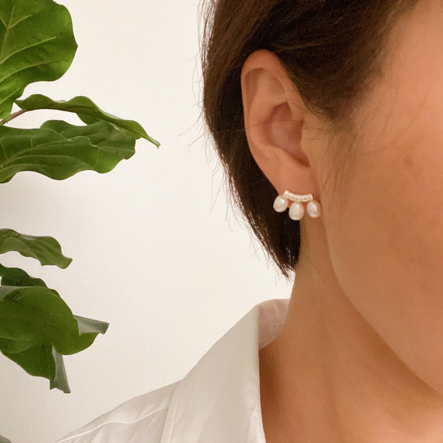 Elegant pearl earrings with diamond accents worn by a woman in a white shirt, surrounded by lush green leaves in a soft, natural setting.