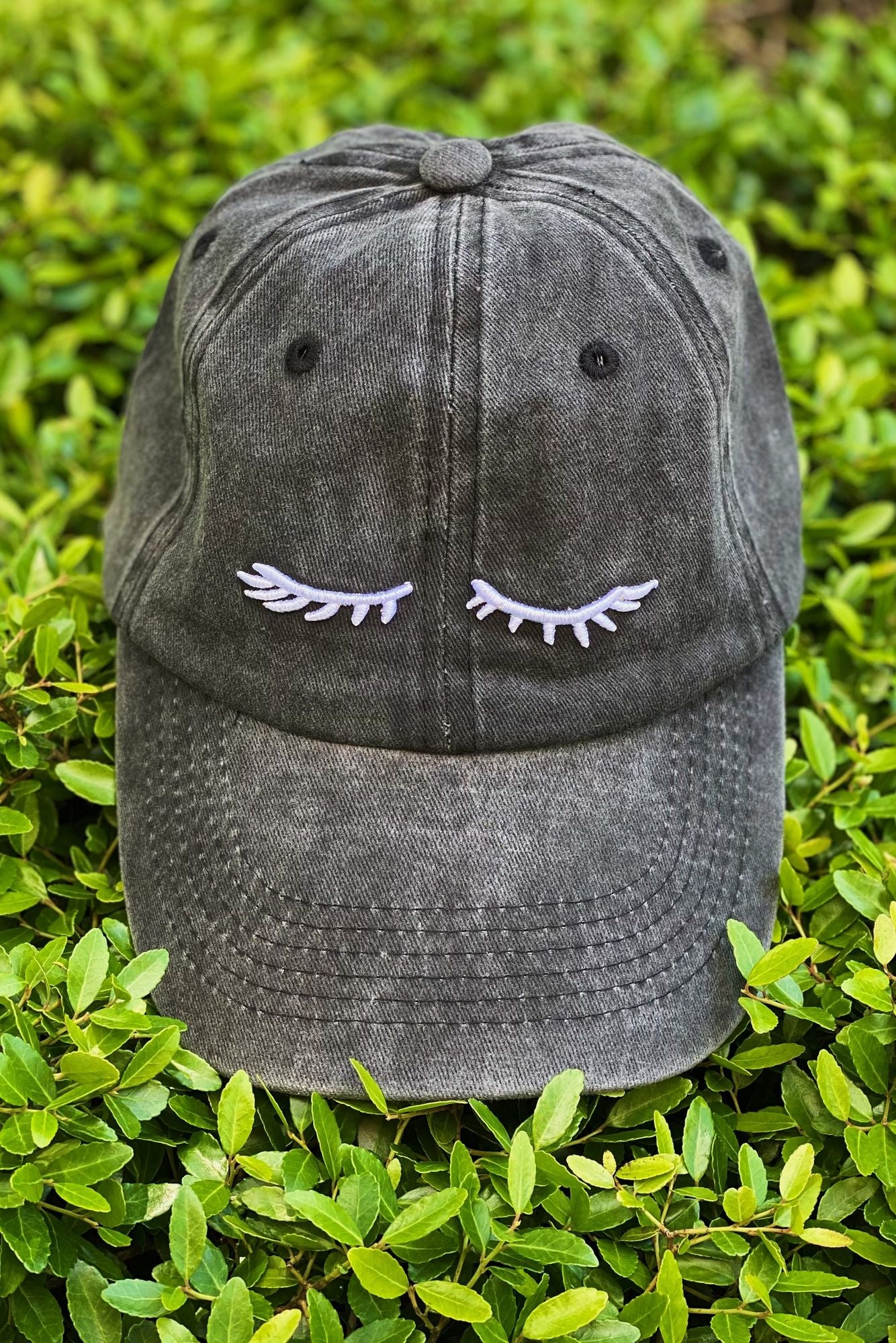 Distressed gray baseball cap with embroidered eyelashes design, resting on a bed of green foliage.