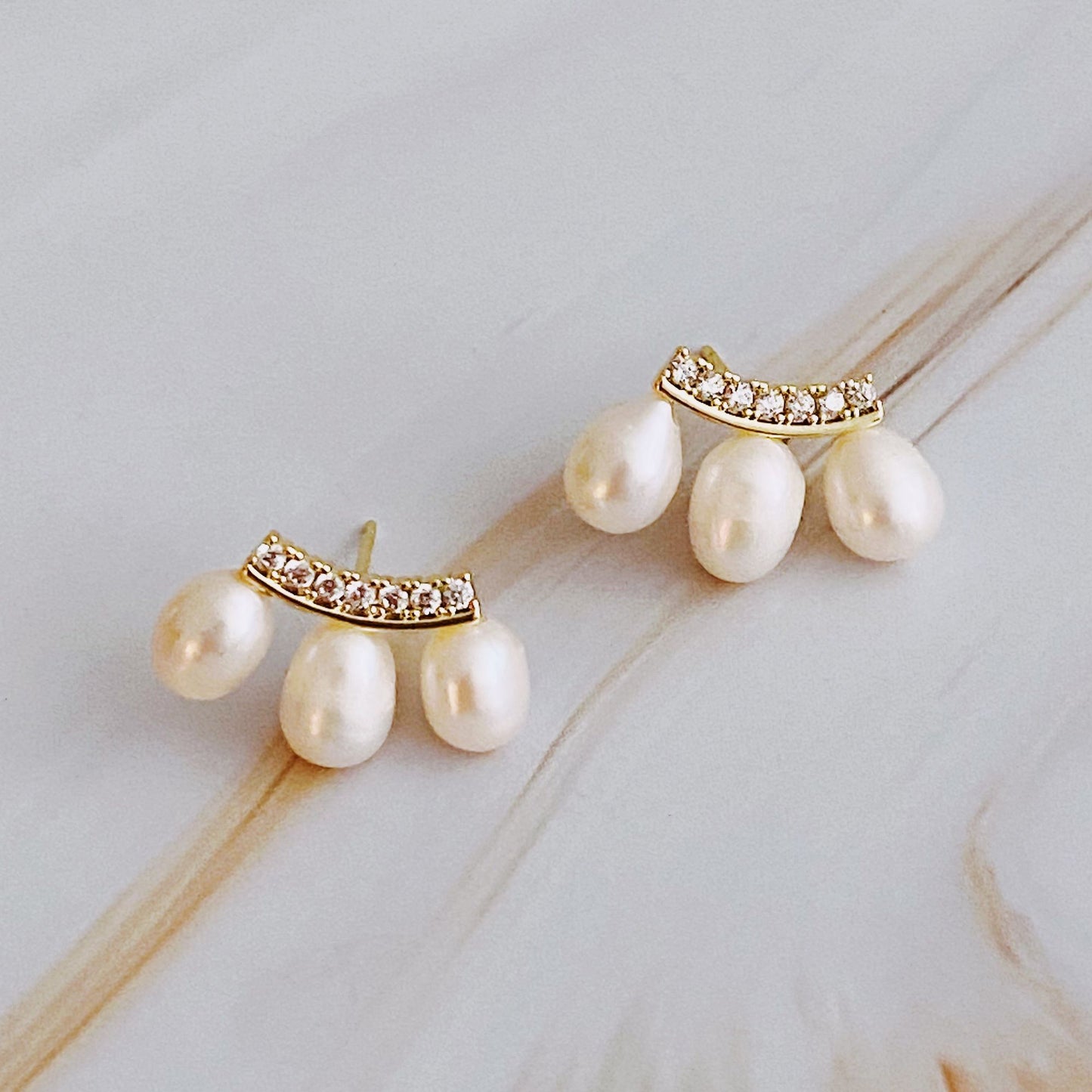 Elegant gold and pearl earrings featuring a curved design adorned with sparkling rhinestones and three dangling freshwater pearls, set against a soft marble background.