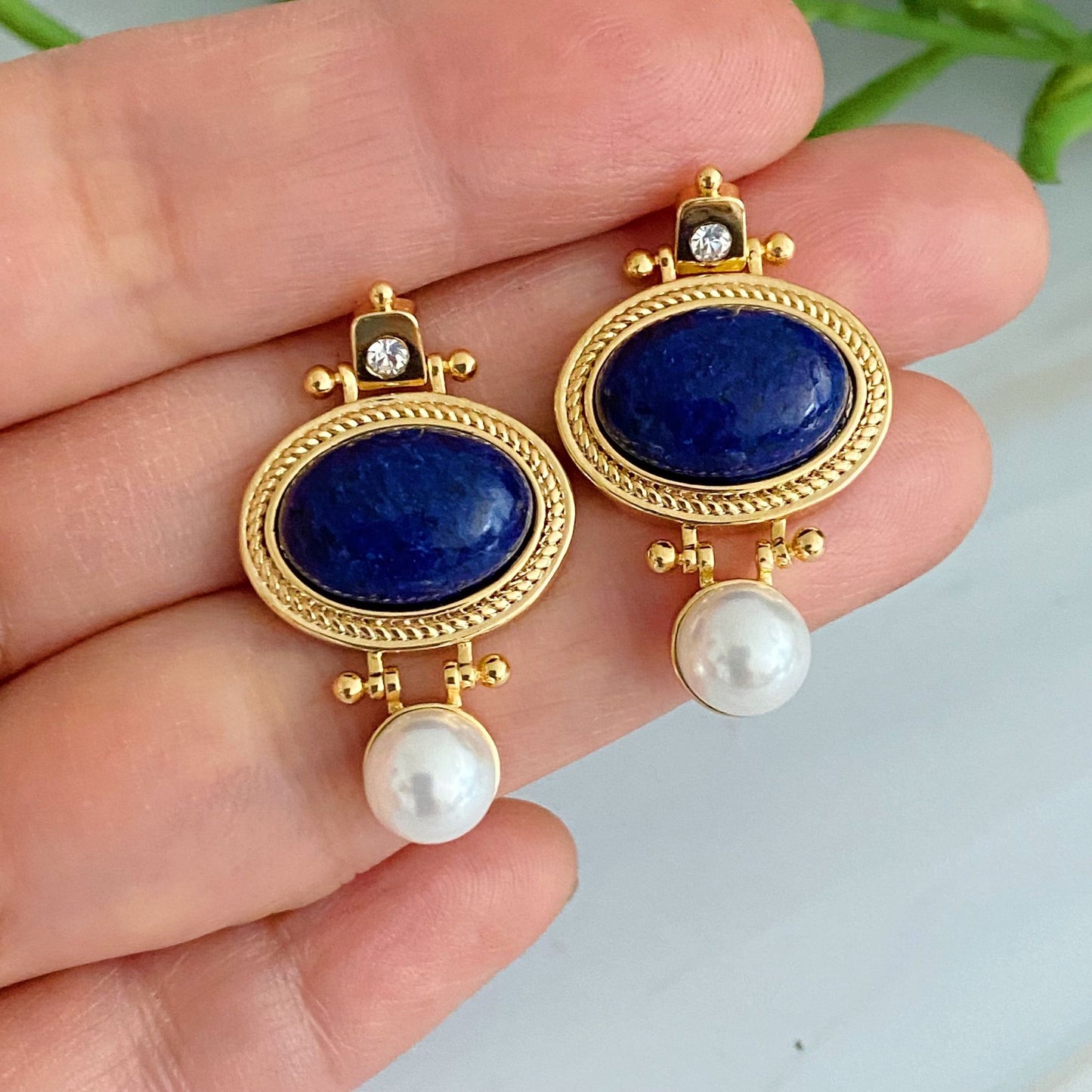 Elegant gold earrings featuring deep blue Lapis Lazuli gemstones, sparkling clear crystals, and lustrous white pearls, displayed on a hand for scale.