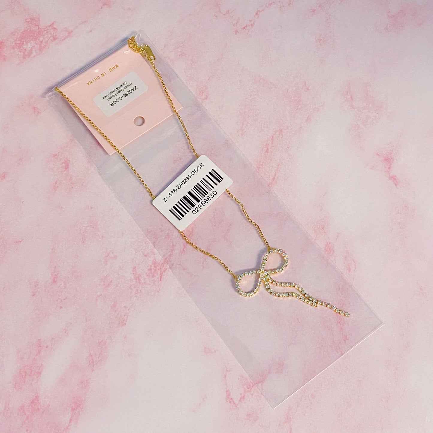 Gold infinity necklace with sparkling rhinestones, presented in clear packaging against a pink marble background.