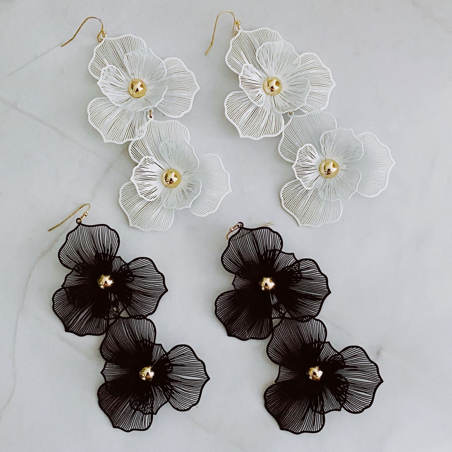 Elegant floral earrings in white and black, featuring intricate layered petal designs with a central gold bead, displayed on a marble background.