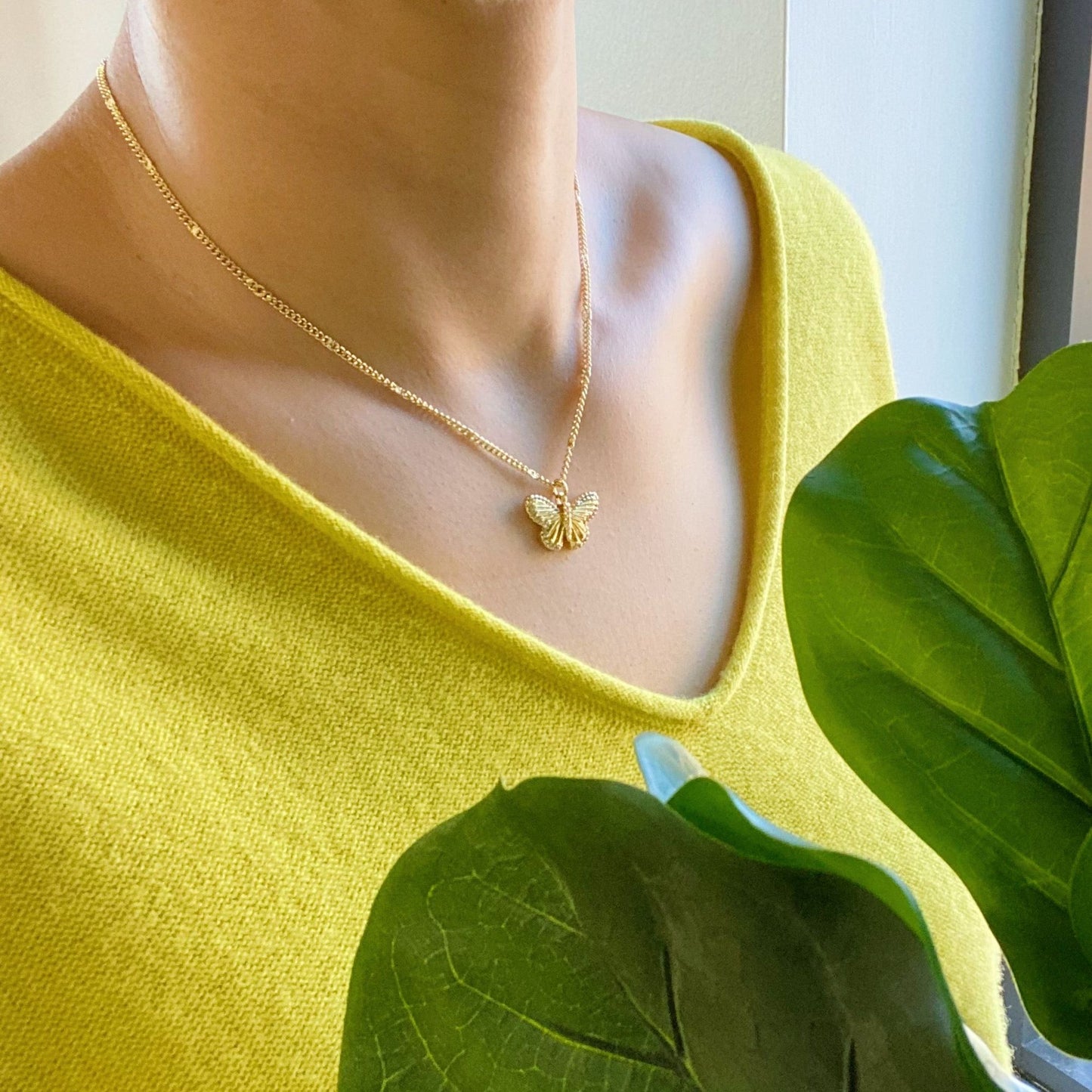 Elegant gold butterfly necklace worn by a woman in a yellow top, accented by lush green leaves in the foreground.