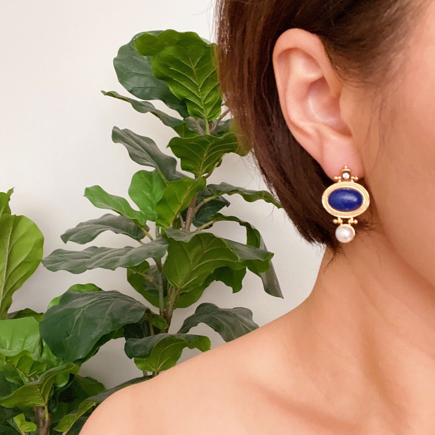 Elegant gold and blue gemstone drop earrings with a pearl accent, showcased on a model against a lush green plant backdrop.