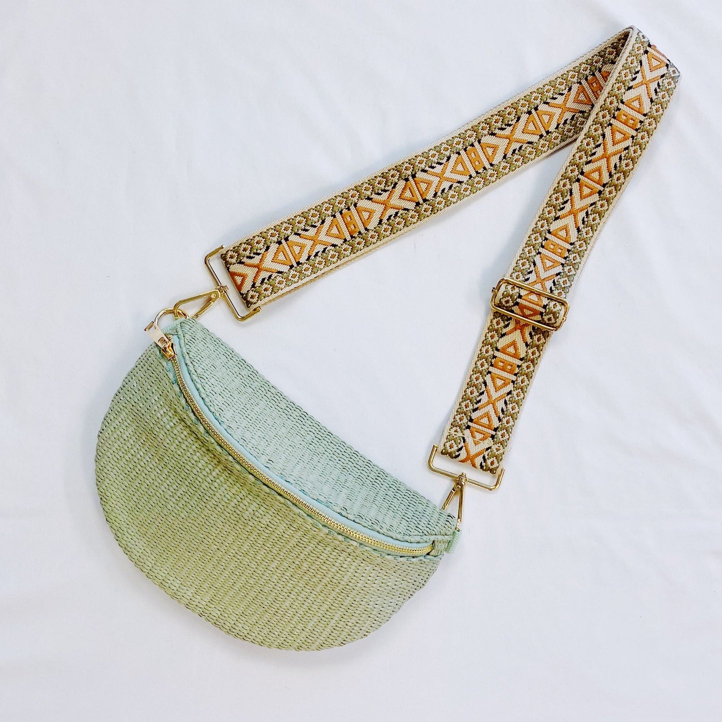 Light green woven crossbody bag with a decorative patterned strap and gold hardware, displayed on a white background.