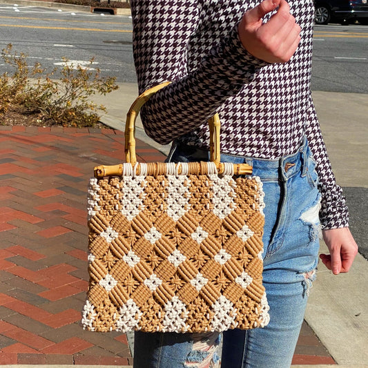 Handwoven tan and white patterned tote bag with bamboo handles, being held by a person in a houndstooth long-sleeve top and distressed denim jeans, outdoors on a sunny day.