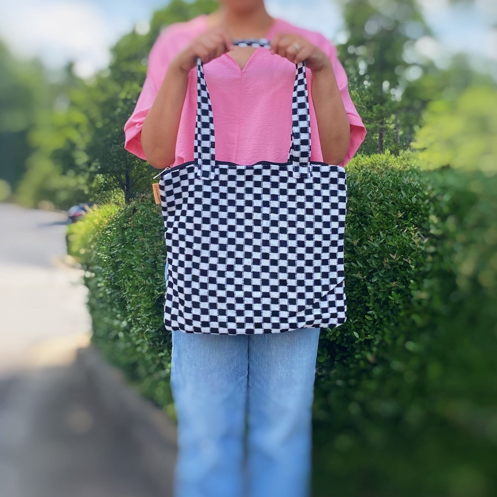 Woman holding a stylish black and white checkered tote bag, wearing a pink top and blue jeans, with a blurred green background.