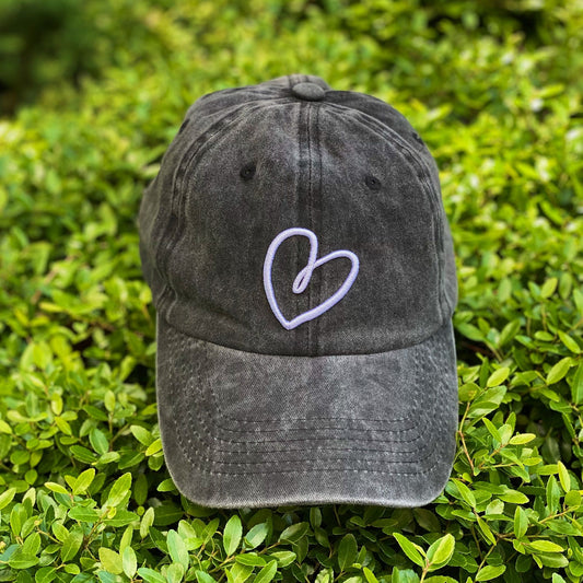 Dark gray baseball cap with white heart embroidery resting on lush green foliage