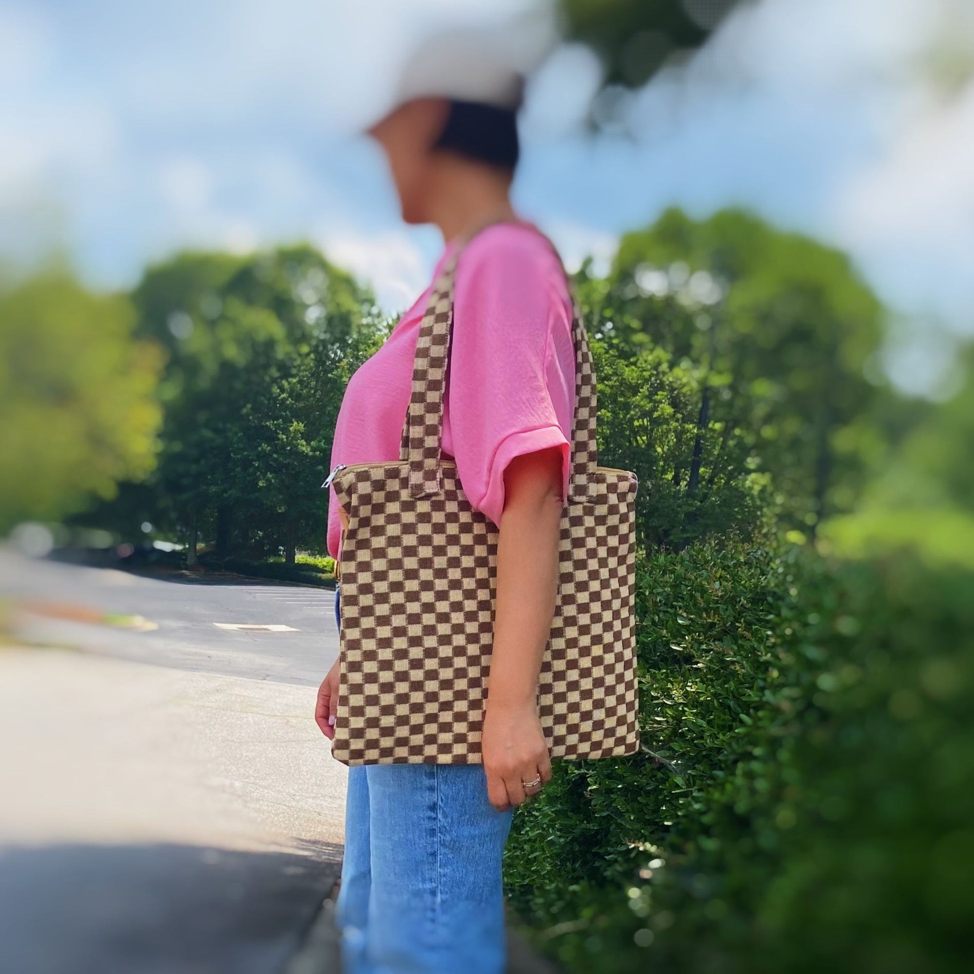 Stylish brown and cream checkered tote bag held by a woman in a pink shirt and jeans, outdoors with greenery in the background.