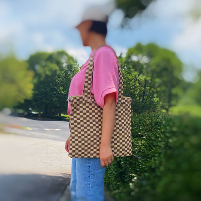 Stylish brown and cream checkered tote bag held by a woman in a pink shirt and jeans, outdoors with greenery in the background.