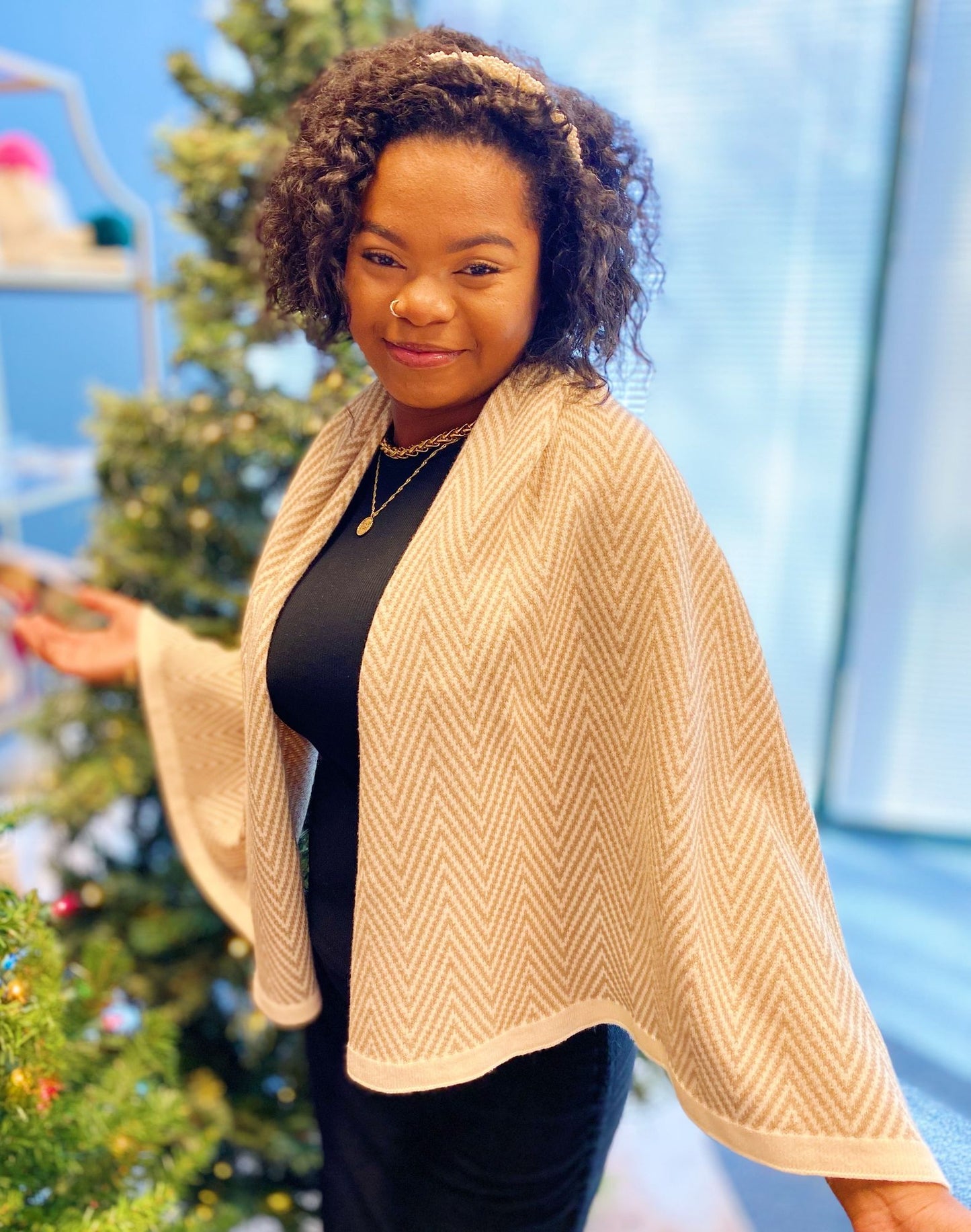 Model wearing a stylish beige herringbone patterned wrap, elegantly draped over a black dress, near a decorated Christmas tree in a bright, festive setting.