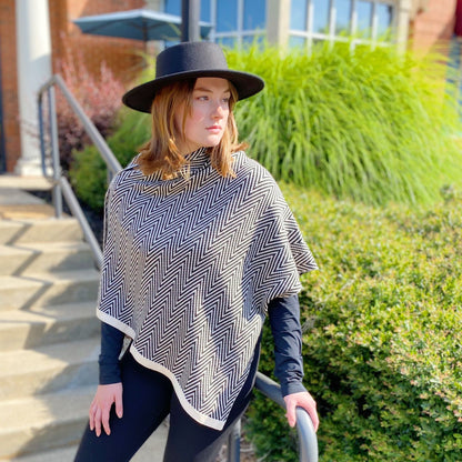 Stylish woman wearing a black and white chevron-patterned poncho and a wide-brimmed hat, standing outdoors near greenery and steps, perfect for a chic fall look.