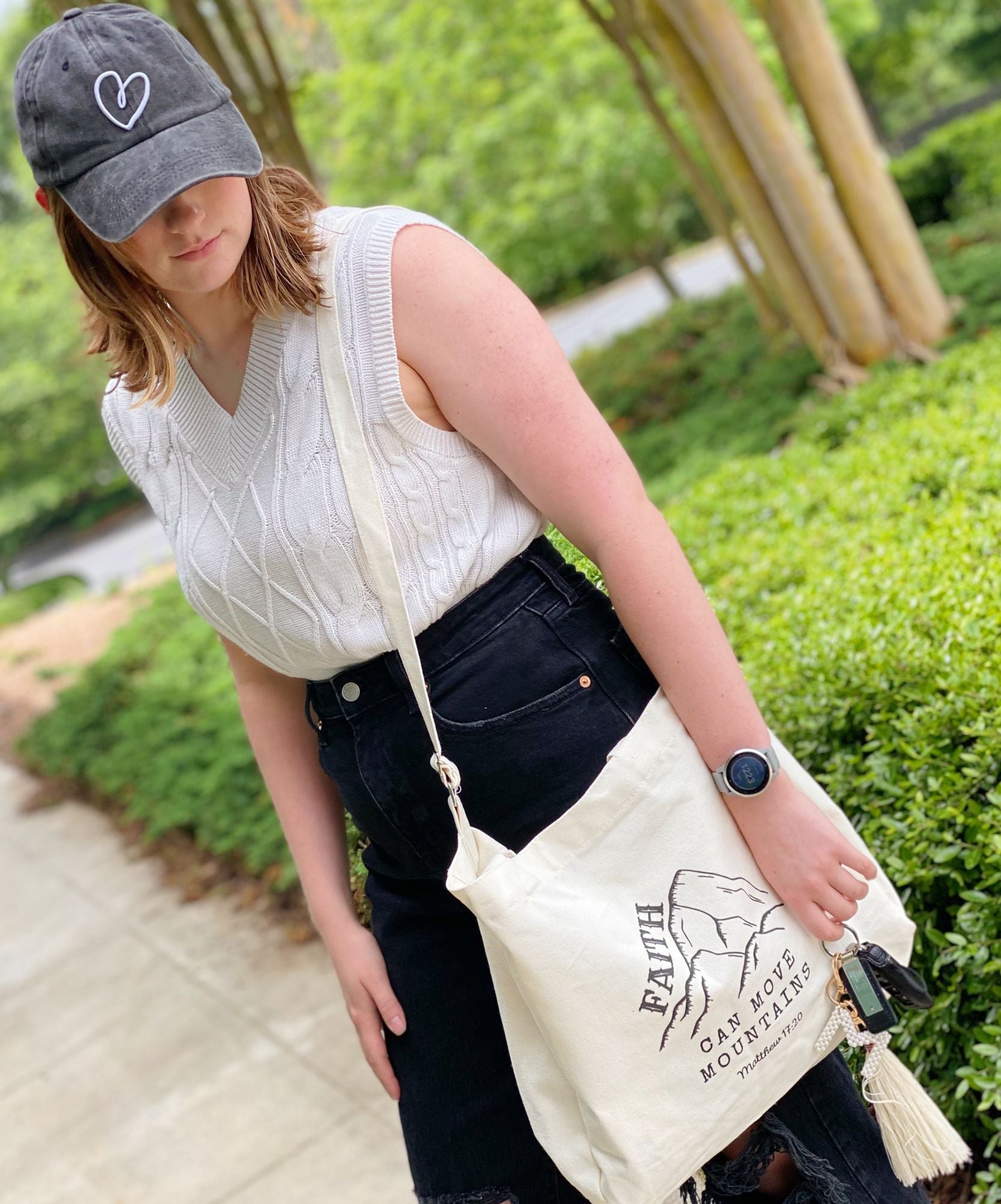 A young woman stands outdoors wearing a stylish white cable-knit sweater and a black denim skirt, holding a cream-colored tote bag featuring the inspirational phrase "Faith Can Move Mountains" and a mountain graphic.