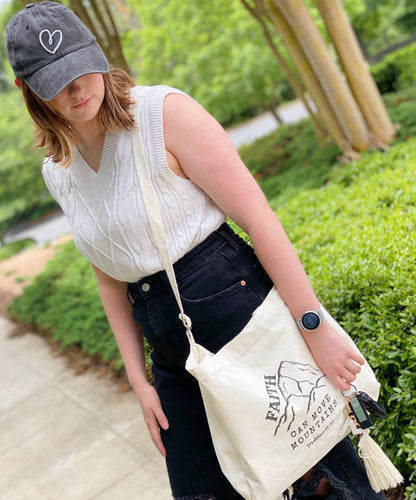 A young woman stands outdoors wearing a stylish white cable-knit sweater and a black denim skirt, holding a cream-colored tote bag featuring the inspirational phrase "Faith Can Move Mountains" and a mountain graphic.