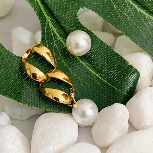 Gold hoop earrings with pearl drops resting on a green leaf atop white decorative stones, showcasing elegant jewelry design.