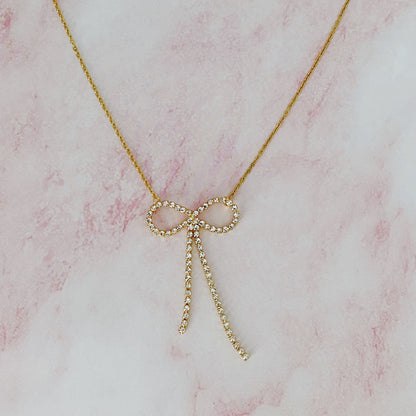 Gold necklace featuring a diamond-studded bow pendant on a delicate chain, set against a soft pink marble background.