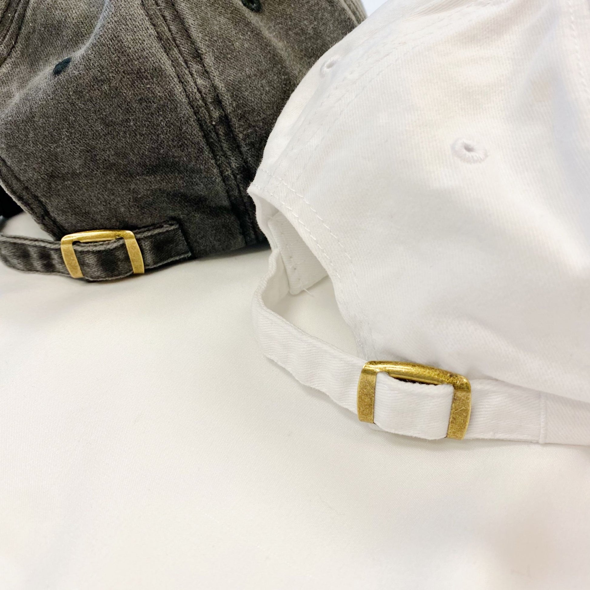 Close-up of a gray and a white baseball cap, showcasing their adjustable straps with gold metal buckles against a neutral background.