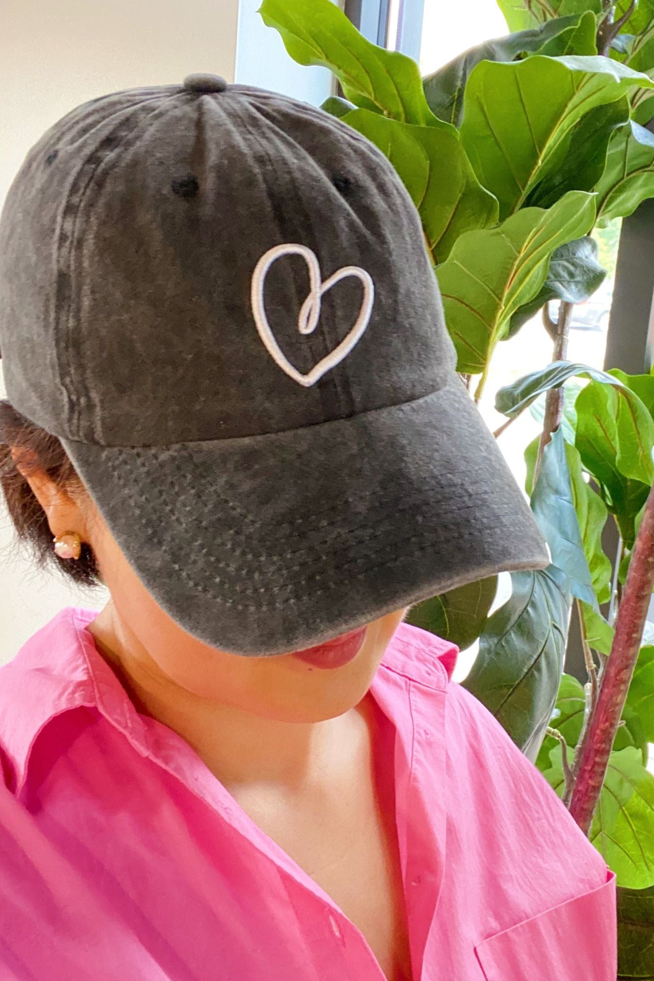 Woman wearing a black baseball cap with a white heart outline design, paired with a pink shirt, in a bright indoor setting with greenery in the background.